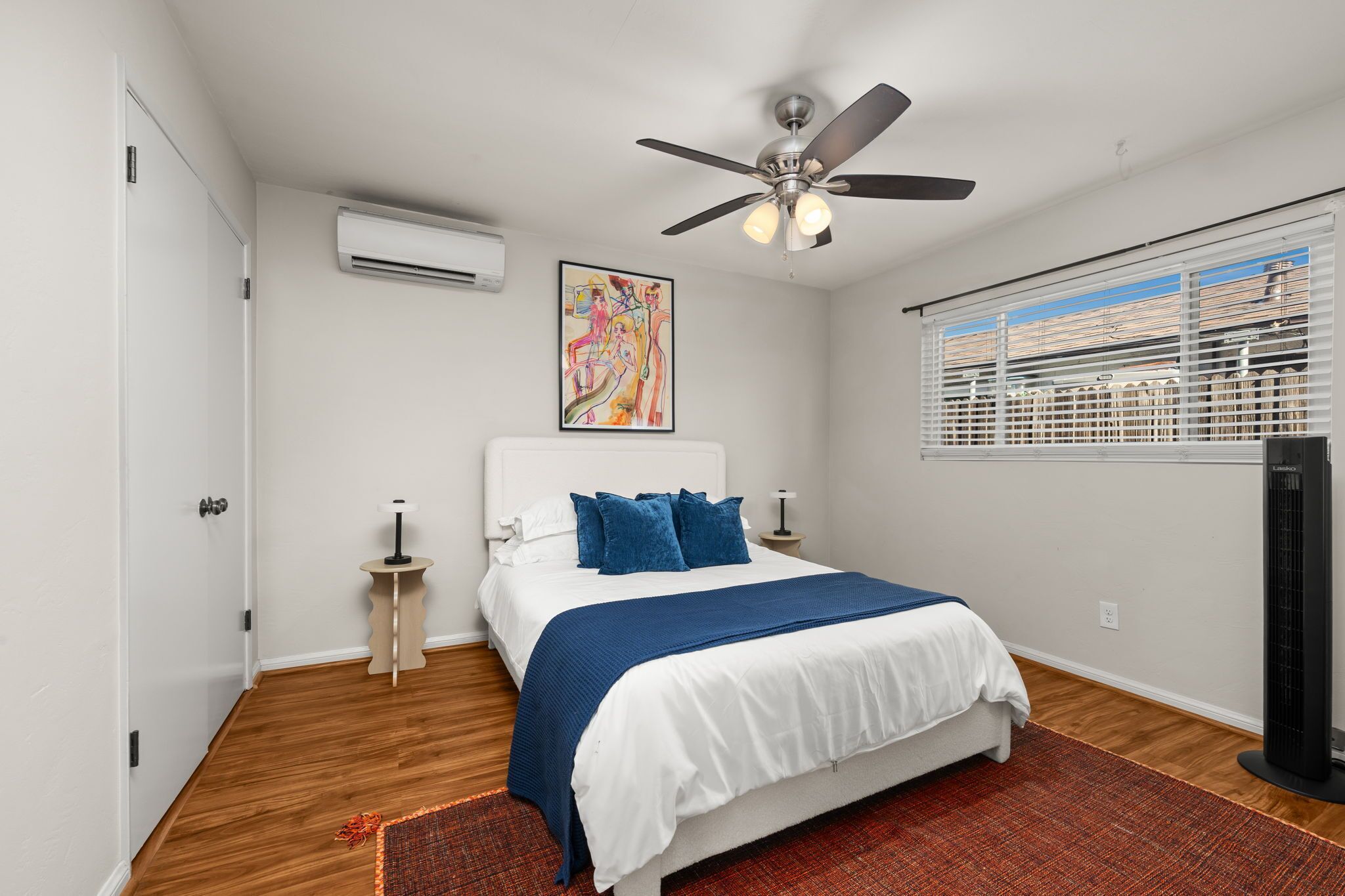 Une chambre à coucher comprenant un lit, un ventilateur de plafond, un appareil de climatisation, des œuvres d’art, des stores et un radiateur mobile.