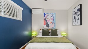 A bedroom with a bed, two bedside lamps, a window with blinds, and two wall art pieces. - Cozy Pacific Beach 2BR Patio Fire Pit (San Diego)