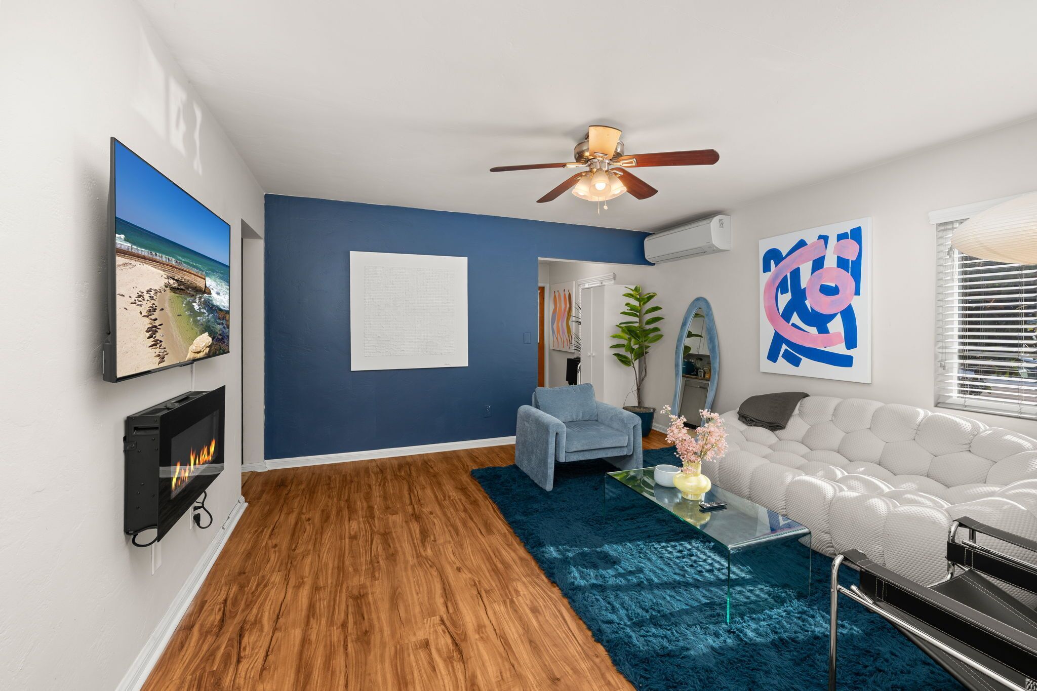 A modern living room with a fireplace, a ceiling fan, a large abstract painting, and a view of the ocean.