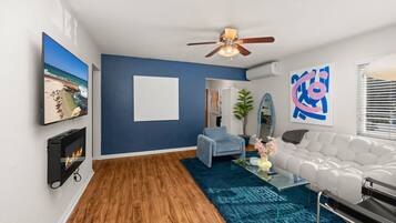 A modern living room with a fireplace, a ceiling fan, a large abstract painting, and a view of the ocean.