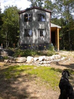 A two-story cabin with a porch, surrounded by a natural landscape. - Cozy, Private 1-bedroom house with stream frontage in tranquil Bridgton with AC (Bridgton)