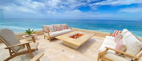 A terrace with a sofa, chairs, and a fire pit overlooking the ocean.
