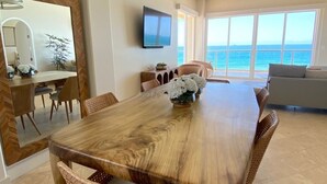 Un espace repas avec une grande table en bois, des chaises en osier, un luminaire suspendu et une vue sur l’océan grâce à de grandes fenêtres.
