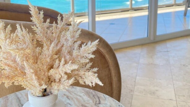 Une pièce avec vue sur l’océan, un vase blanc contenant des plantes séchées et une table en marbre.