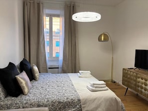 A hotel room with a bed, a TV, a floor lamp, and a view of buildings outside. - Cozy apartment in the Vatican district (Roma)
