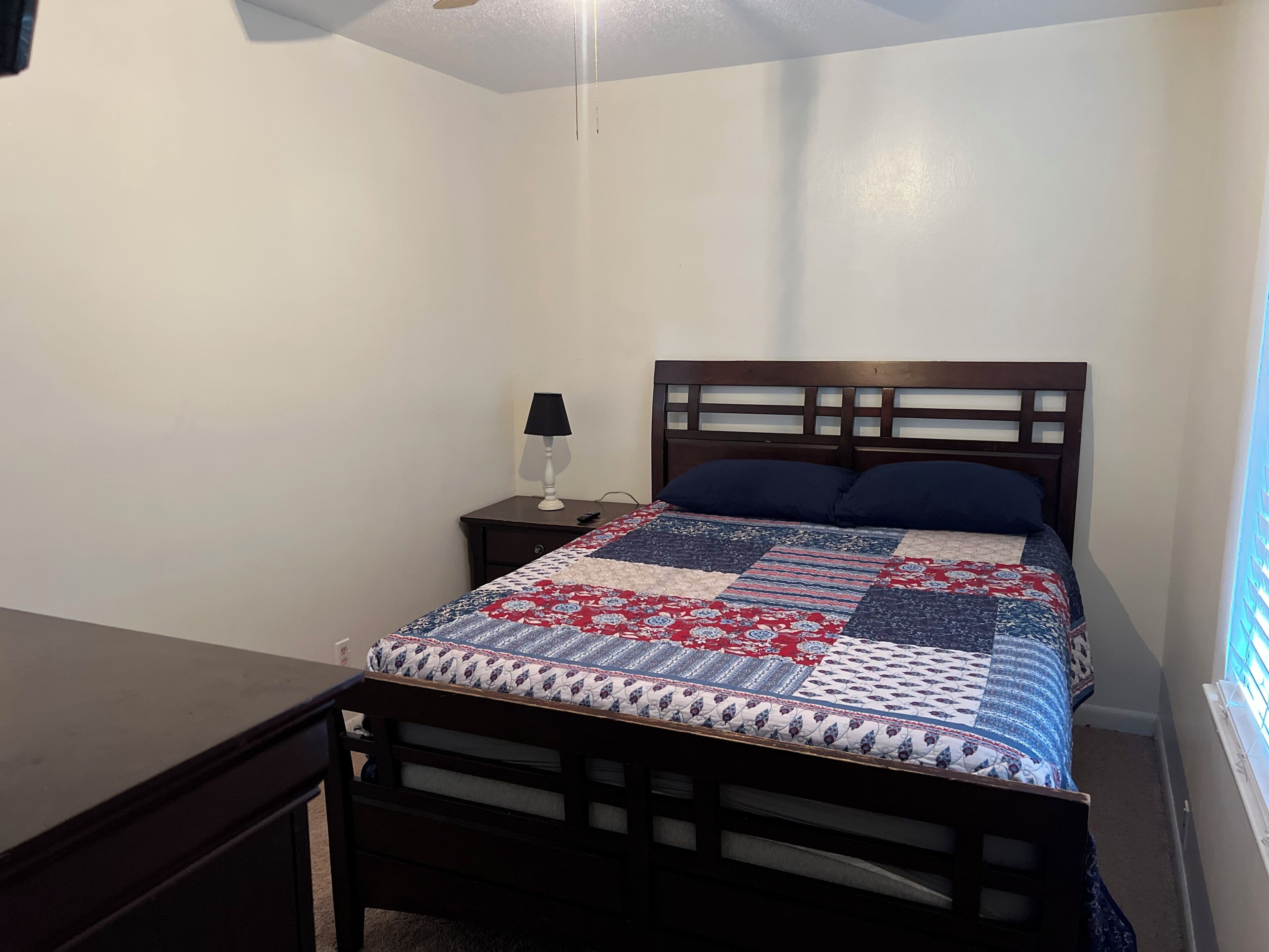 Een slaapkamer met een houten bed, een nachtkastje, een lamp en een raam met jaloezieën.