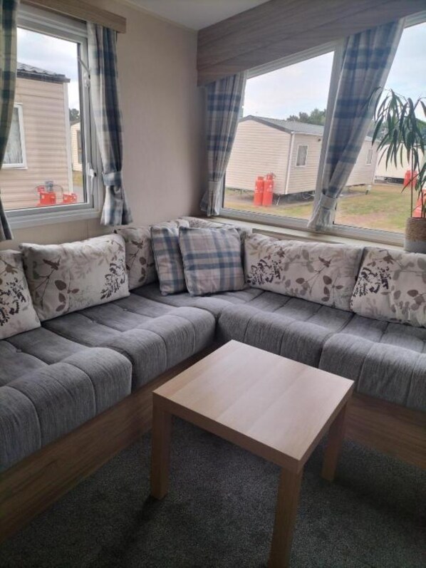 A sectional sofa with floral cushions, a wooden coffee table, and large windows with curtains. - Lovely 3-bedroom caravan in quaint Port Seton perfect for relaxing (Port Seton)