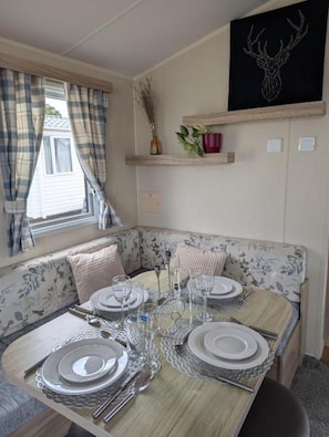 A dining area with a table set for four, a floral sofa, a window with curtains, and a wall-mounted shelf with decorative items.