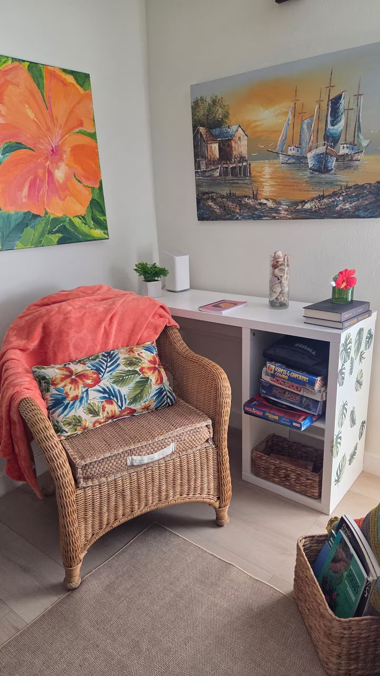 Une pièce chaleureuse avec un fauteuil en osier, un bureau avec des jeux de société et deux tableaux accrochés au mur.