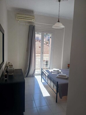 Ein Schlafzimmer mit einem Bett, einem kleinen Tisch mit Teekanne, einem Fenster mit Vorhängen und einer Klimaanlage.