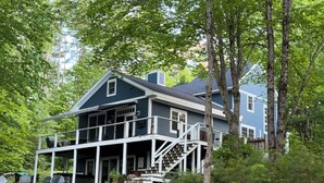 A two-story house with a porch, a staircase leading to the upper floor, and a surrounding wooded area. - Welcome to the Bear’s Den. A Delightful house on Lake Winnipesaukee in Meredith. (Meredith)