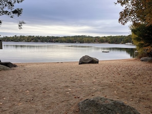 A lakeside view with a sandy beach, large rocks, and a distant shoreline.