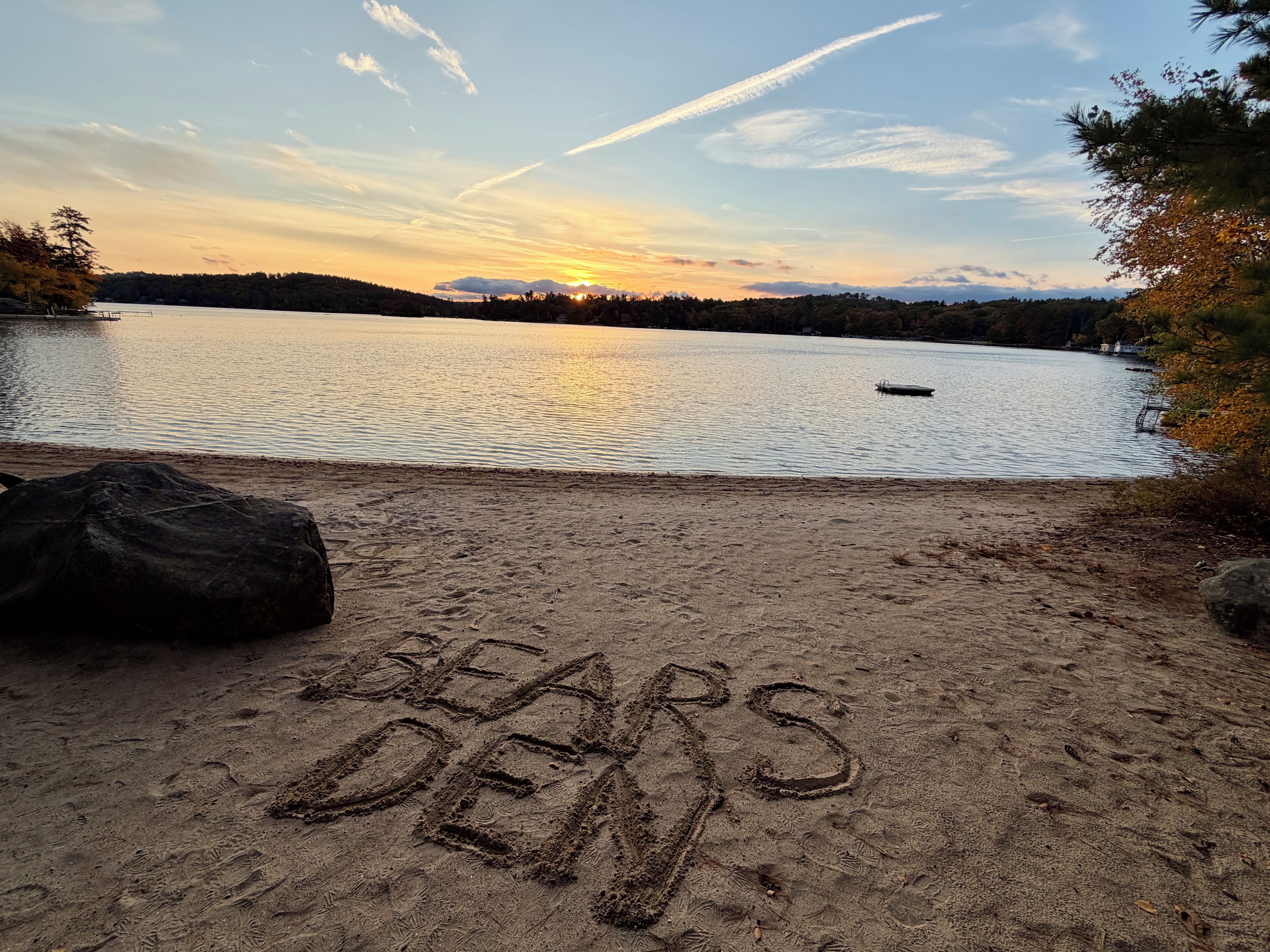 A beach with the words 'BEARS DEN' written in the sand, a calm lake, and a sunset in the background.
