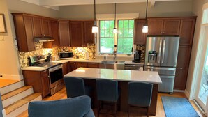 A modern kitchen with wooden cabinets, a central island with bar stools, stainless steel appliances, and a view of greenery through large windows. - Welcome to the Bear’s Den. A Delightful house on Lake Winnipesaukee in Meredith. (Meredith)