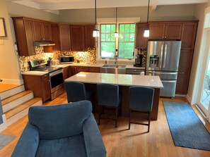 A modern kitchen with wooden cabinets, a central island with bar stools, stainless steel appliances, and a view of greenery through large windows.