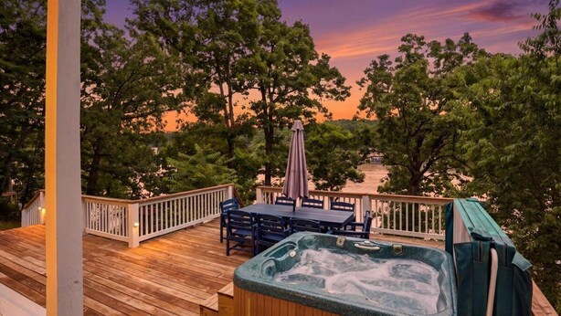 A wooden deck with a hot tub, outdoor furniture, and a view of trees and a sunset.