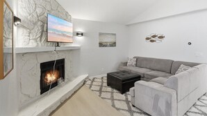 A living room with a fireplace, a gray sofa, a black ottoman, and a TV mounted on a stone wall.