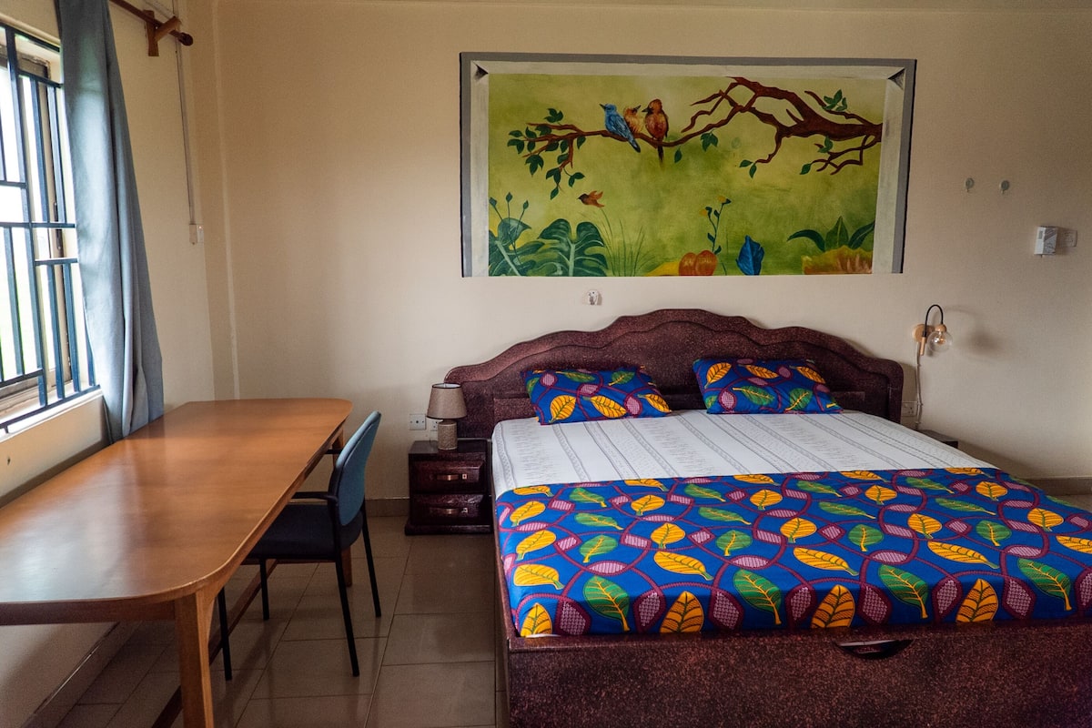 A bedroom with a bed, a desk, and a chair. There is a window with bars and a curtain.