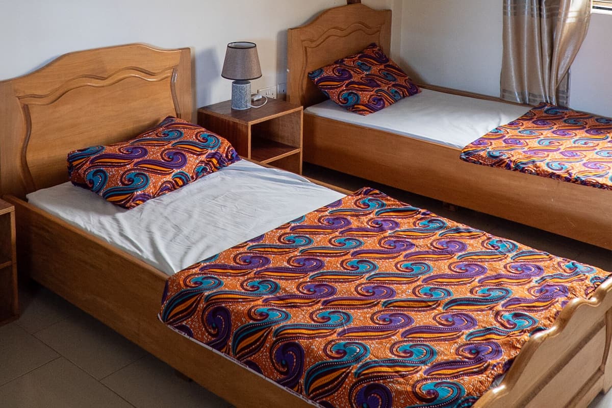 A room with two wooden beds, each with patterned bedding, a nightstand with a lamp, and a wall mural of a tree with hanging leaves.
