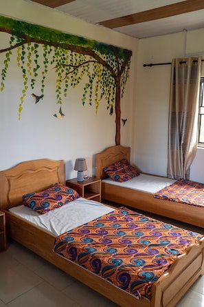 Una habitación con dos camas de madera, cada una con ropa de cama estampada, una mesita de noche con lámpara y un mural en la pared de un árbol con hojas colgantes.
