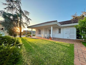 A white house with a tiled roof, a covered patio with a table and chairs, and a well-maintained lawn. - Casa Sierrapando - by Wehomes (Torrelavega)