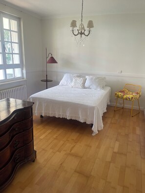 A bedroom with a large bed, a dresser, a chair, a chandelier, and a window. - Casa Sierrapando - by Wehomes (Torrelavega)