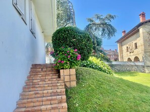 Un escalier mène à un bâtiment blanc dont la base est en briques, entouré d’une végétation bien entretenue et d’un mur de pierre.