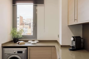 Een kleine keuken met een wasmachine, een koffieapparaat, een bloempot en uitzicht op gebouwen door een raam.