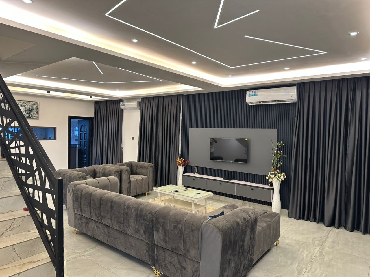 A modern living room with a sectional sofa, a coffee table, a flat-screen TV, and a staircase.