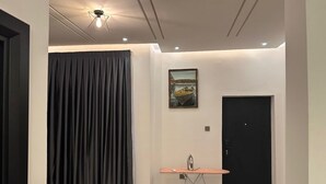 Una habitación con puerta, ventana con cortinas, una mesa con plancha y un cuadro en la pared.