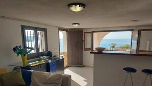 Een woonkamer met een bank, een salontafel, een bureau met een glazen blad en uitzicht op zee door een groot raam.
