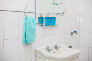 Ein Badezimmer mit einem weißen Waschbecken, zwei Wasserhähnen, einem Handtuchhalter mit einem blauen Handtuch und zwei blauen Flaschen auf einem Glastischchen über dem Waschbecken.