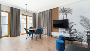 Una sala de estar moderna con piso de madera, una mesa de comedor con sillas azules, un televisor de pantalla plana montado en un panel de madera y un ventanal con cortinas.