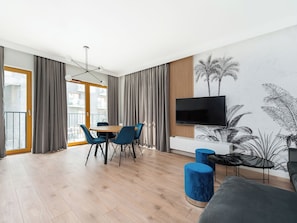 A modern living room with a wooden floor, a dining table with blue chairs, a flat-screen TV mounted on a wooden panel, and a large window with curtains. - Apartment Novum by Interhome (Krakow)