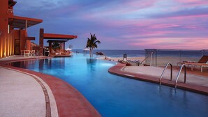 Une piscine avec vue sur la plage et l’océan, entourée de chaises longues et d’un espace abrité.