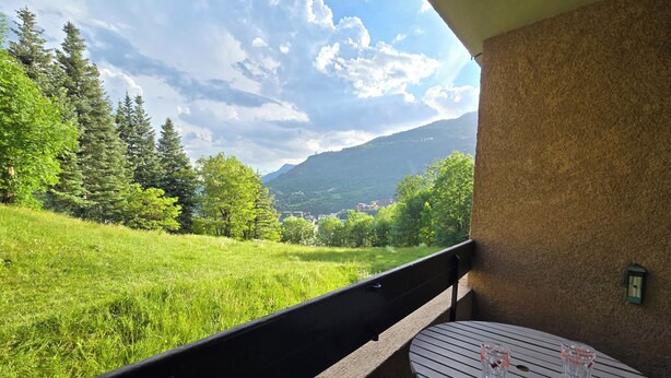 A balcony with a table and two glasses, overlooking a grassy field and a mountain. - South-facing mountain apartment: summer/winter ideal for biking, climbing, hiking and skiing (Briançon)