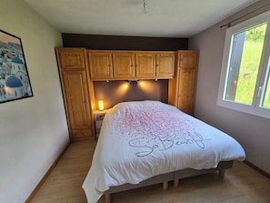 Un dormitorio con cama, armarios de madera, mesita de noche, lámpara, ventana con vistas y un cuadro en la pared.