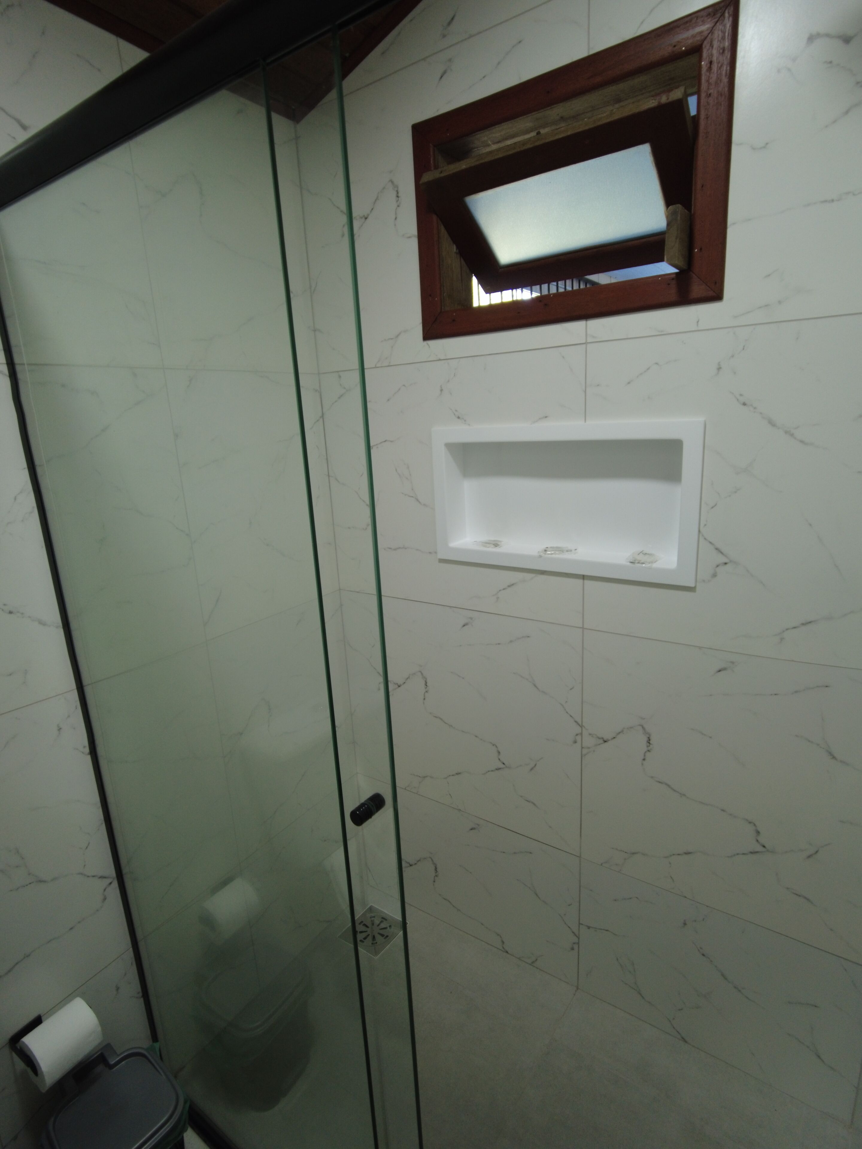 A bathroom with a glass shower enclosure, a marble-tiled wall, a small shelf, and a toilet with a paper towel holder.