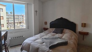 Ein Schlafzimmer mit einem Bett, einem Fenster, einem Kruzifix an der Wand und Blick auf Gebäude.