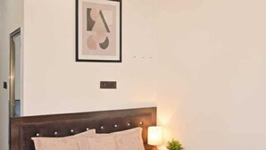A bedroom with a bed, a nightstand with a lamp, a framed picture on the wall, and a ceiling light fixture. - Monishka Palace  (Faizabad)