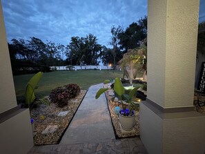 Un sendero conduce a un jardín con un estanque, rodeado de follaje y una valla blanca.