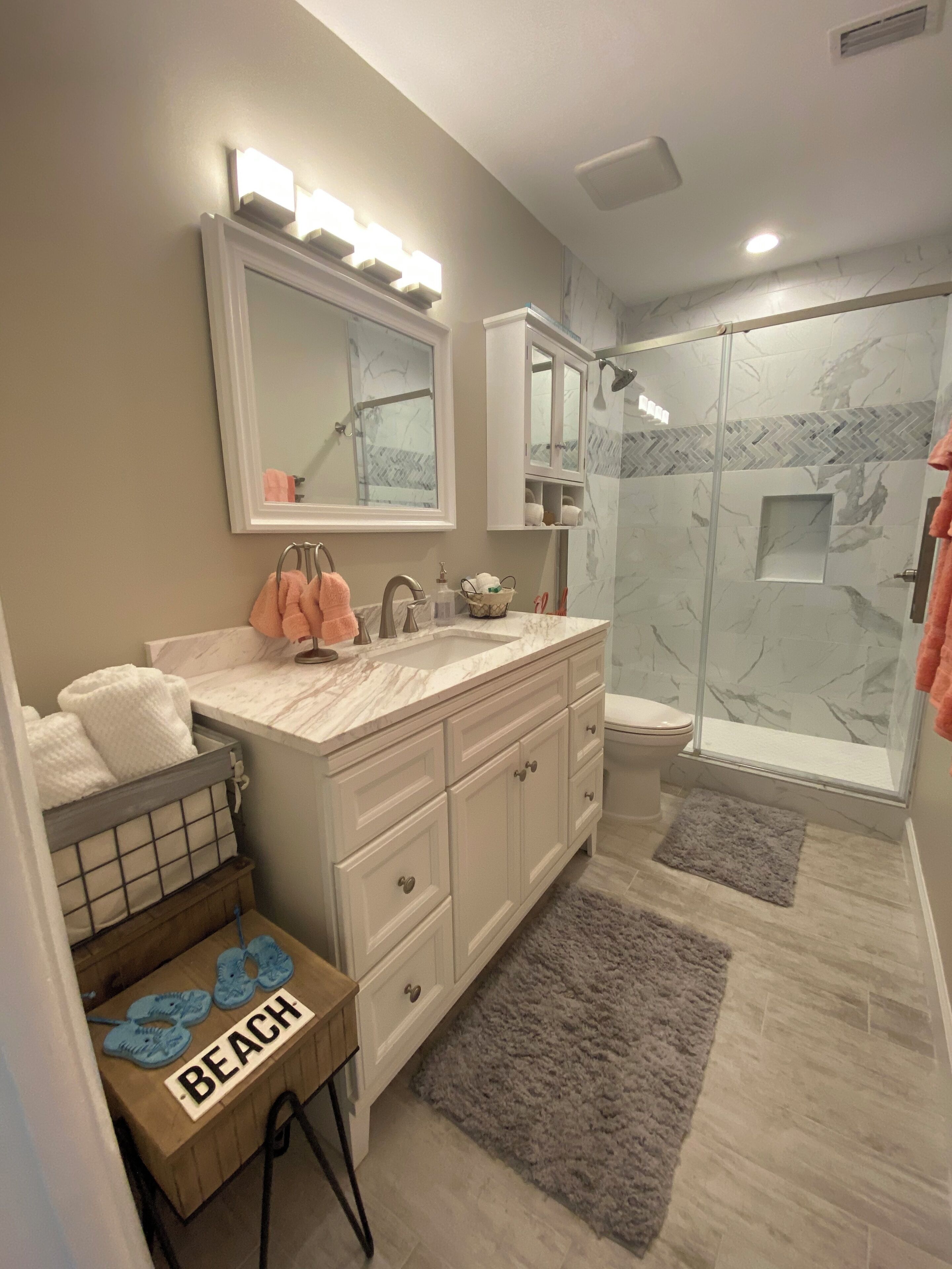 A bathroom with a vanity, a shower, and a small table with a beach-themed sign.