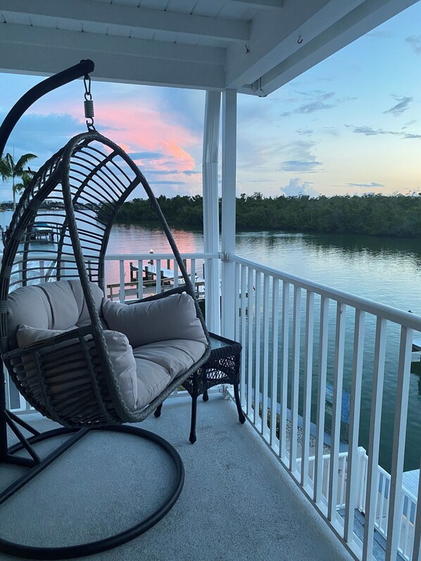 A balcony with a hanging chair, a small table, and a view of a body of water with a dock and trees in the background. - Beautiful Waterfront Condo on Old Marco (Marco Island)