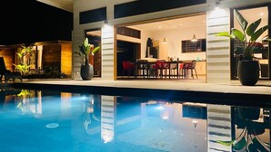 A swimming pool with a modern house in the background, a dining area with red chairs, and potted plants. - Villa Te Nunoa - Polynesian Villa w/ Salt Water Pool (Haapiti)