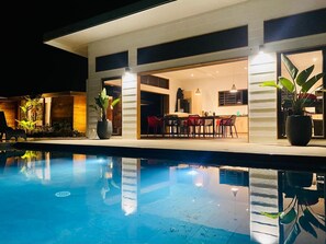 Ein Schwimmbecken mit einem modernen Haus im Hintergrund, einem Essbereich mit roten Stühlen und Topfpflanzen.