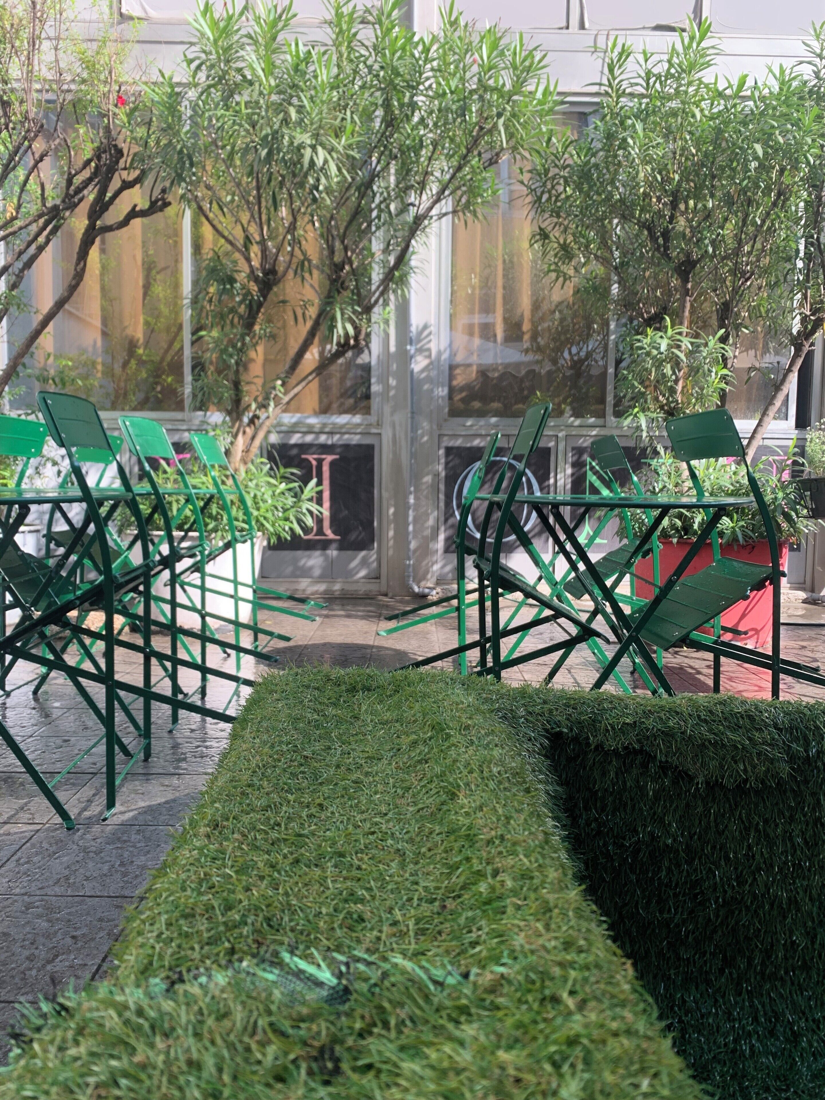 Un espace extérieur aménagé avec des chaises et des tables vertes, entouré de haies soigneusement taillées et de plantes en pot.