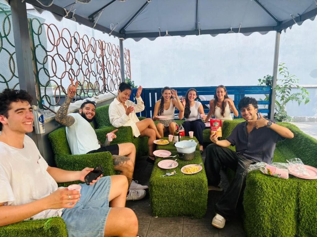 Un groupe de personnes est assis sur des meubles de jardin en gazon synthétique vert, sous une tonnelle. Un mur décoratif en métal se trouve à l’arrière-plan.