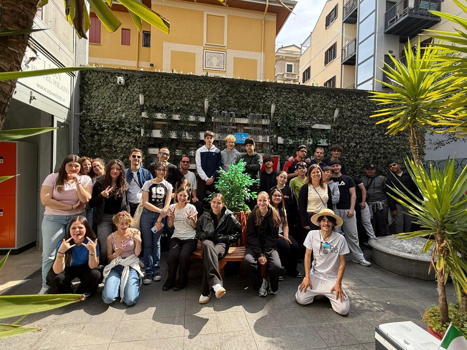 Un gruppo di persone riunito in uno spazio all'aperto con un muro in pietra e piante, di fronte a un edificio con un cartello che recita 'LIONHOSTEL.IT'.
