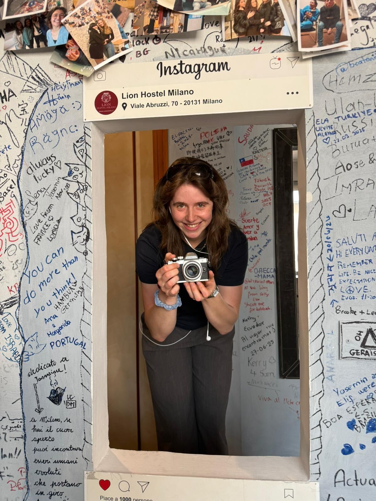 Une personne prend une photo à l’intérieur d’une pièce dont les murs sont couverts de notes et de dessins manuscrits, où se trouve un miroir et un panneau indiquant l’auberge de jeunesse Lion Hostel Milano.