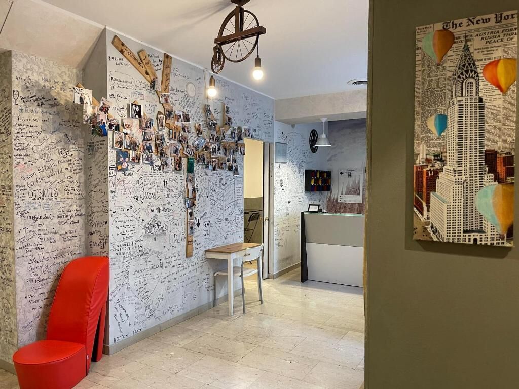 Une pièce dont un mur est recouvert de notes manuscrites, avec une chaise rouge, un bureau et un tableau représentant une vue de la ville.